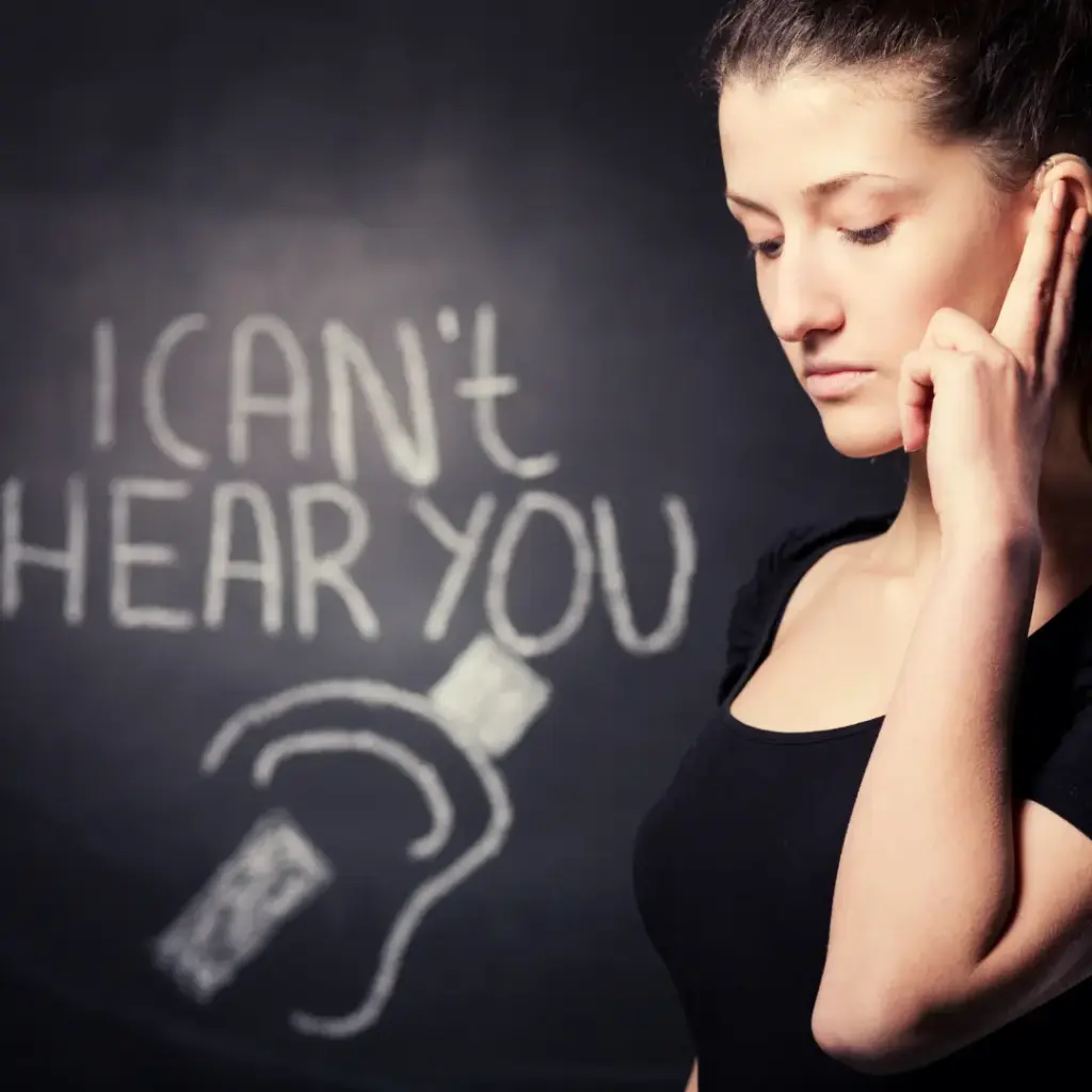 A woman holds her ear and stands before a blackboard that says, “I can’t hear you.”