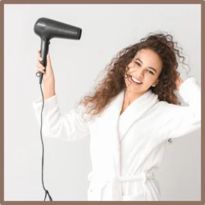A woman in a bathrobe points her hair dryer at her ear. 