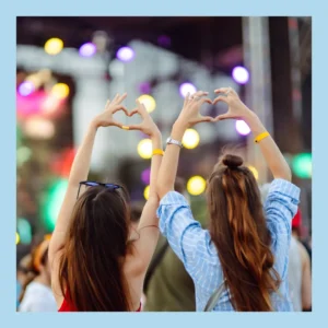 Women stand far from the stage at an outdoor concert and hold their hands in heart shapes. 