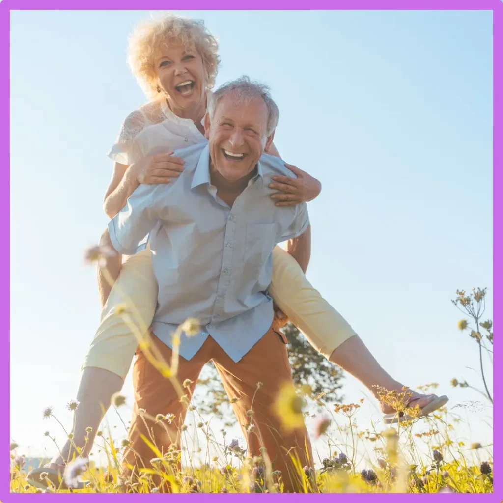 An older man gives an older woman a piggyback ride in a field.