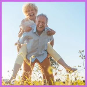 An older man gives an older woman a piggyback ride in a field.