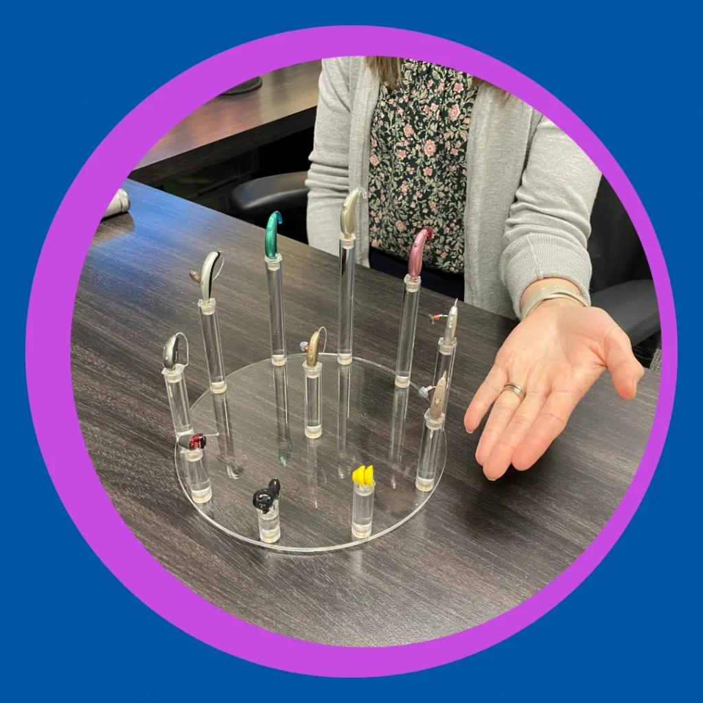 A hand points to a tray of hearing aid models.